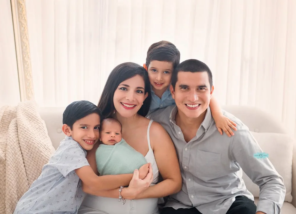 familia de cinco con ropa coordinada en tonos azul suave y gris, abrazando a su recién nacido durante una sesión de fotos familiar